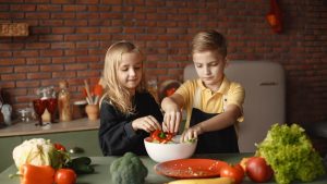 Cheerful girl helping little boy in apron preparing salad while standing in kitchen near table with mix of fresh vegetables behind brick wall